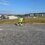 A construction worker in high-visibility clothing uses a GPS surveying instrument on a gravel site with buildings and grassy fields in the background under a clear sky.