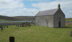 A small stone church stands beside a graveyard on grassy land, with hills and a body of water in the background under a partly cloudy sky.