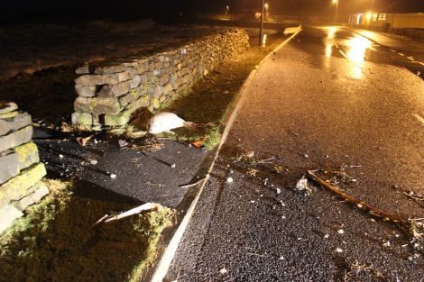 The grey seal pup shelters behind a wall opposite Lerwick fire station with waves crashing ashore send ing spray across the road.