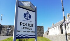 A sign outside a stone building reads "Police Scotland, Police Lerwick, Shetland Area Office" on a clear day with a blue sky.