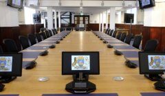 A large, rectangular conference table with black chairs and individual monitors displaying a crest, located in a well-lit room with several wall-mounted screens and a stained glass window in the background.