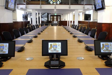 A large, rectangular conference table with black chairs and individual monitors displaying a crest, located in a well-lit room with several wall-mounted screens and a stained glass window in the background.