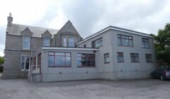 A grey stone building with a modern extension, surrounded by a paved area and flanked by a small car parked on the right side. The sky is overcast.