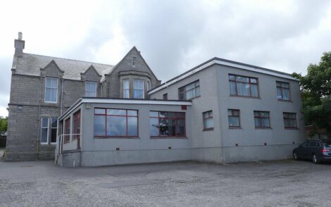 A grey stone building with a modern extension, surrounded by a paved area and flanked by a small car parked on the right side. The sky is overcast.