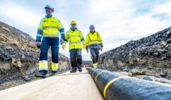 Three workers in high-visibility clothing and helmets walk on a wooden path next to a large underground cable in a trench at a construction site.