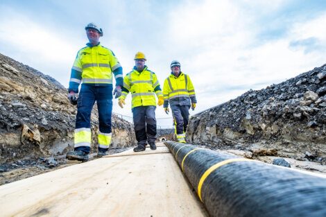 Three workers in high-visibility clothing and helmets walk on a wooden path next to a large underground cable in a trench at a construction site.