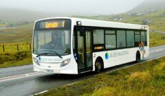 A white bus with "LERWICK E" displayed on the front LED screen is driving on a wet countryside road, with misty hills and scattered houses in the background.