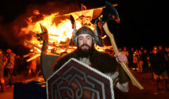 A man in Viking attire holds an axe and shield in front of a large bonfire at night, with a crowd of people in the background.