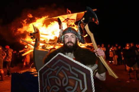 A man in Viking attire holds an axe and shield in front of a large bonfire at night, with a crowd of people in the background.