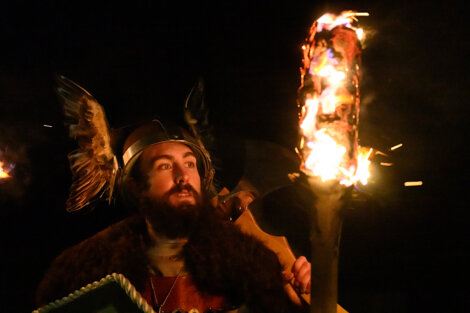 A person wearing a Viking helmet with feathers, holding a burning torch with a focused expression, against a dark background.