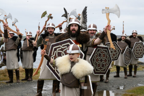 A group dressed in Viking attire, complete with helmets, shields, and axes, pose spiritedly outdoors near a body of water.