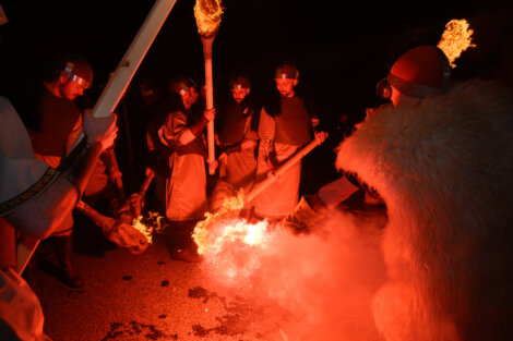 People in costumes holding torches gather around a fire at night, with one person wearing a fur cloak and a red helmet.