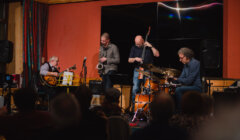 A four-member jazz band performs on stage with guitar, saxophone, double bass, and drums, in front of an audience.