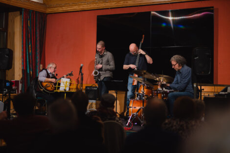 A four-member jazz band performs on stage with guitar, saxophone, double bass, and drums, in front of an audience.