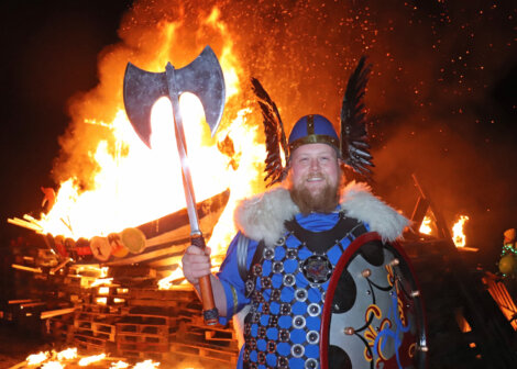 A man in a Viking costume stands in front of a large bonfire, holding a double-bladed axe and a decorated shield, with a burning Viking ship in the background.