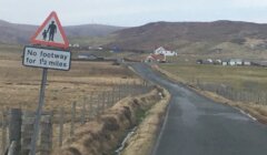 Rural road with a sign: "No footway for 1½ miles." The road is flanked by fences, leading towards distant houses and hills.