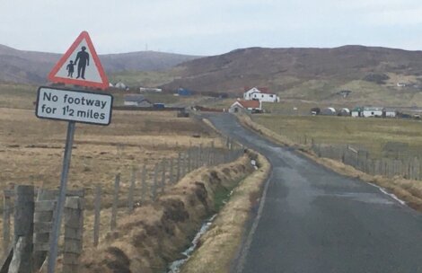 Rural road with a sign: "No footway for 1½ miles." The road is flanked by fences, leading towards distant houses and hills.