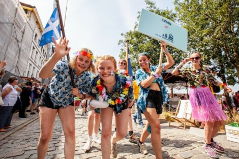 Children wearing Hawaiian-themed outfits and flower garlands cheer at an outdoor event. One child holds a sign reading "SWAN UK" with a crowd watching in the background.