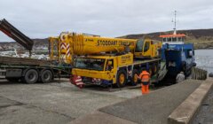 A large yellow crane truck marked "Stoddart Crane Hire" is driven off a blue ferry ramp onto a dock, assisted by workers in orange and reflective uniforms. A hilly landscape is visible in the background.