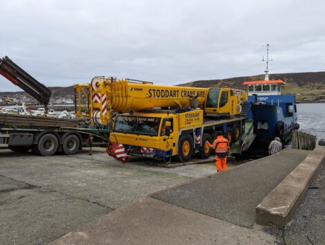 A large yellow crane truck marked "Stoddart Crane Hire" is driven off a blue ferry ramp onto a dock, assisted by workers in orange and reflective uniforms. A hilly landscape is visible in the background.