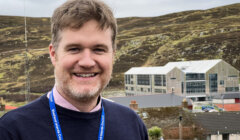 A man with a badge stands smiling outdoors, with a hilly landscape and buildings in the background.