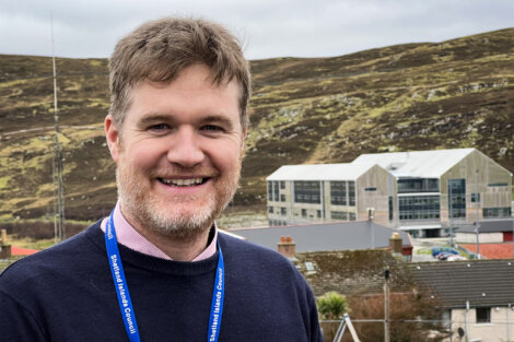 A man with a badge stands smiling outdoors, with a hilly landscape and buildings in the background.