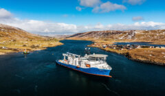 An aerial view of a large blue and white ship navigating a narrow, winding waterway flanked by hills and scattered houses under a partly cloudy sky.