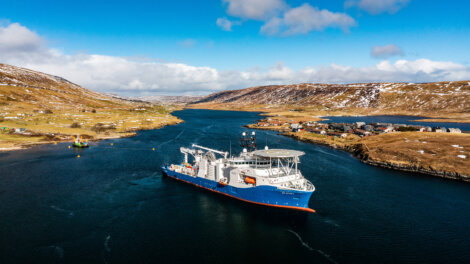 An aerial view of a large blue and white ship navigating a narrow, winding waterway flanked by hills and scattered houses under a partly cloudy sky.