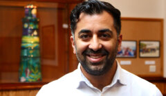 A man with a beard and short hair smiles while standing indoors near colorful artwork and framed photos displayed on a wall behind him.