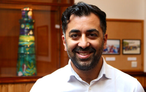 A man with a beard and short hair smiles while standing indoors near colorful artwork and framed photos displayed on a wall behind him.