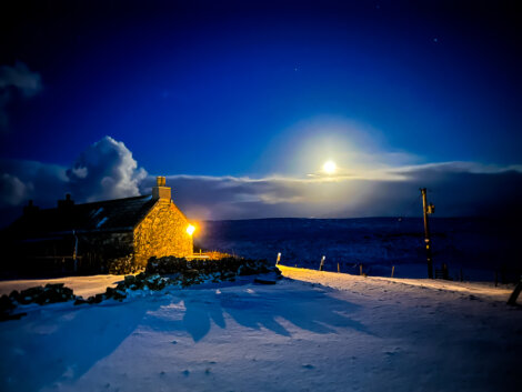 A stone house with a glowing window stands in a snow-covered landscape at night, illuminated under a bright moon and clear sky.