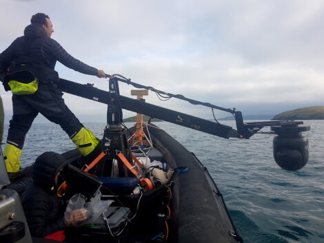 A person stands on a boat operating a camera crane system extended over the water, with equipment and another person visible on the boat.