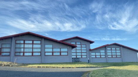 A building with red trim and multiple windows stands under a blue sky with scattered wispy clouds. The structure has three main sections and is surrounded by grass and a paved area.