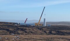 A construction site for wind turbines with cranes lifting large cylindrical components in a remote, hilly area under a clear sky.