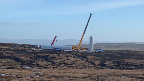 A construction site for wind turbines with cranes lifting large cylindrical components in a remote, hilly area under a clear sky.