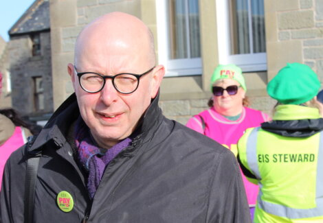 A man with glasses and a bald head, wearing a black jacket with a "Pay Attention" badge, stands outdoors near two people in bright vests and hats marked "EIS Steward.