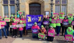 A group of people stand outside holding signs that read “PAY ATTENTION” and “OFFICIAL PICKET” with a banner that says "eis Shetland Local Association." They are protesting in front of a building.