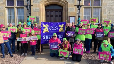 A group of people stand outside holding signs that read “PAY ATTENTION” and “OFFICIAL PICKET” with a banner that says "eis Shetland Local Association." They are protesting in front of a building.