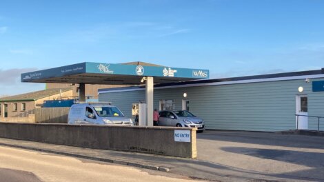 A drive-through COVID-19 testing site with a canopy, signboards, and two vehicles present. One person is visible near the building. A "No Entry" sign is on a wall in front.