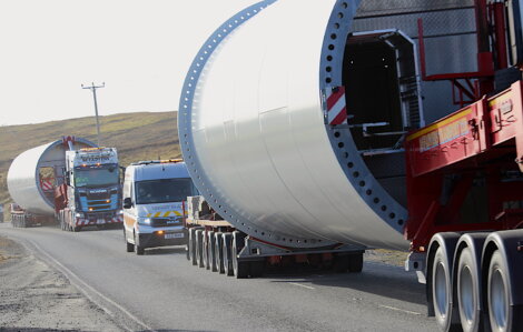 Three trucks transporting large cylindrical structures on a road in daylight, with a hilly backdrop.