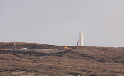 A wind turbine construction site in a landscape with brown and rocky terrain. Machinery and a partially erected wind turbine tower are visible.