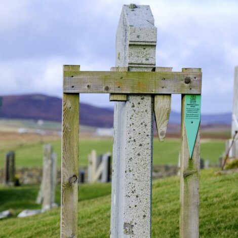 A tilted gravestone supported by wooden braces in a hillside cemetery, with a backdrop of rolling fields and distant hills.