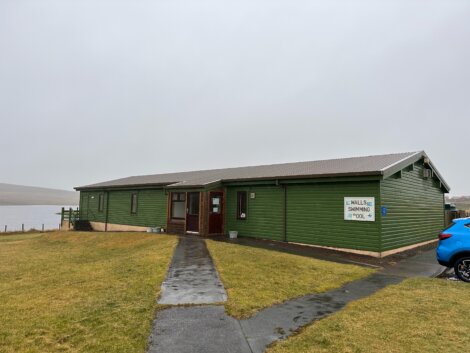 A green building labeled "Swimming Pool" with a sloped roof stands beside a grassy area on a cloudy day. A blue car is parked nearby.