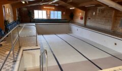 An empty indoor swimming pool with tiled walls, metal handrails, and a viewing gallery sign. The pool area has wooden ceilings and walls, and chairs are lined up on the left side.