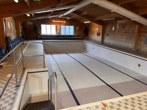 An empty indoor swimming pool with tiled walls, metal handrails, and a viewing gallery sign. The pool area has wooden ceilings and walls, and chairs are lined up on the left side.