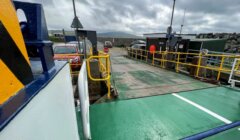 A ferry ramp prepares to dock, with vehicles waiting at the terminal under an overcast sky. The scene includes barriers, safety signs, and a small building.