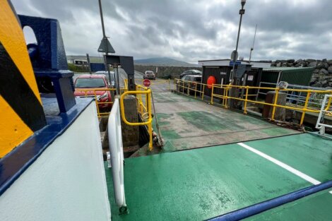 A ferry ramp prepares to dock, with vehicles waiting at the terminal under an overcast sky. The scene includes barriers, safety signs, and a small building.