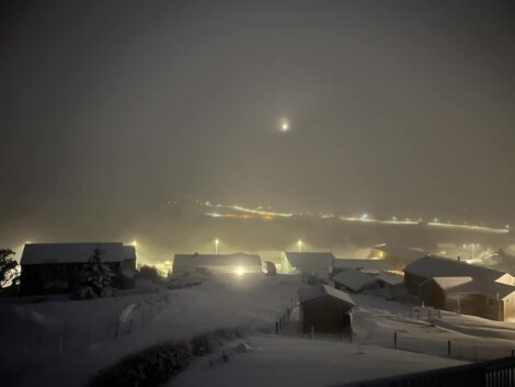 Snow-covered town at night with glowing streetlights and a faintly visible moon in the foggy sky.