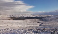 Snow-covered landscape with rolling hills under a partly cloudy sky. A body of water is visible in the distance.