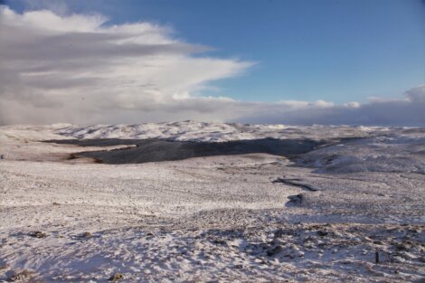 Snow-covered landscape with rolling hills under a partly cloudy sky. A body of water is visible in the distance.